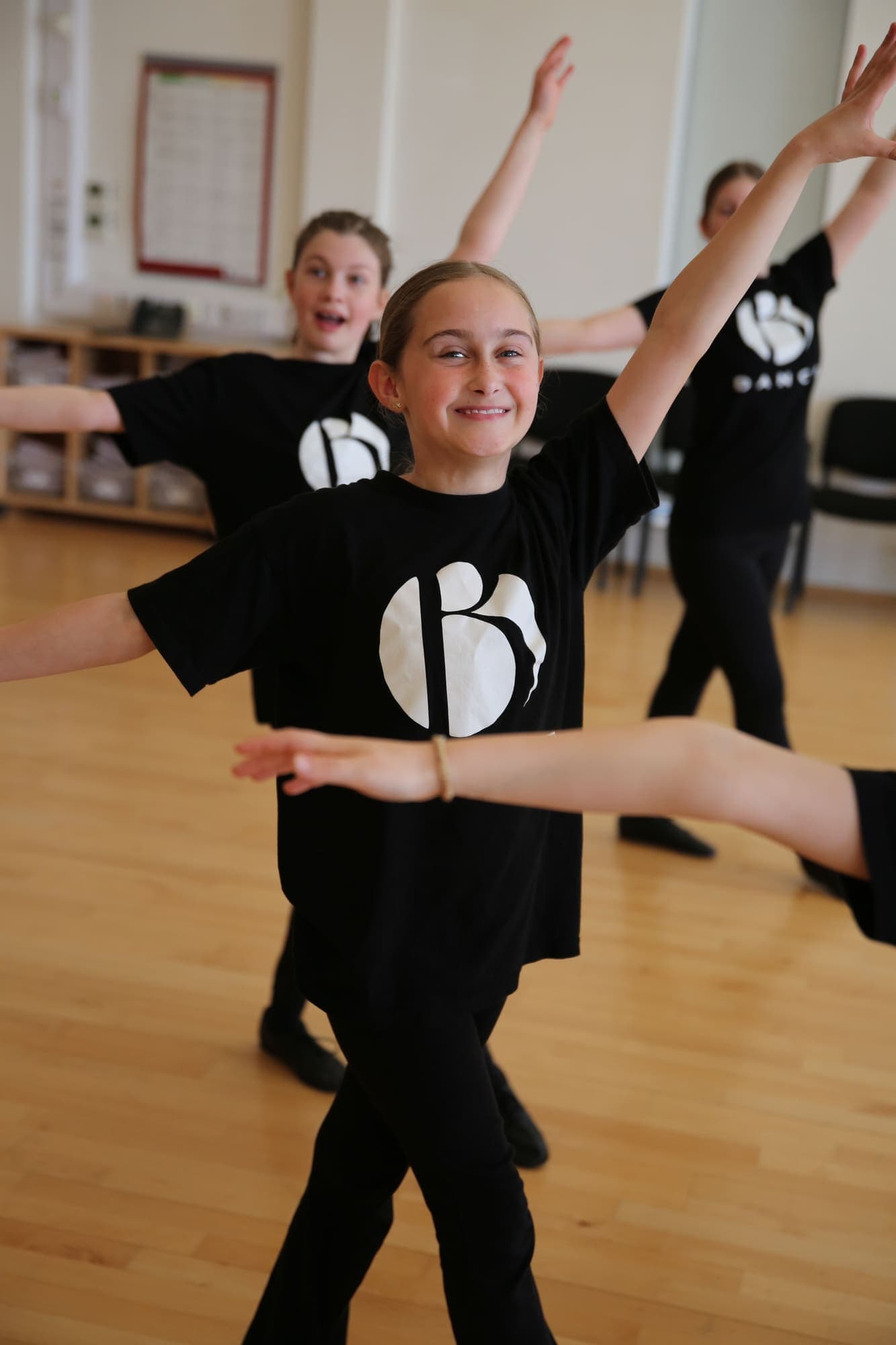 A grinning dancer with arms outstretched, the class moving behind her